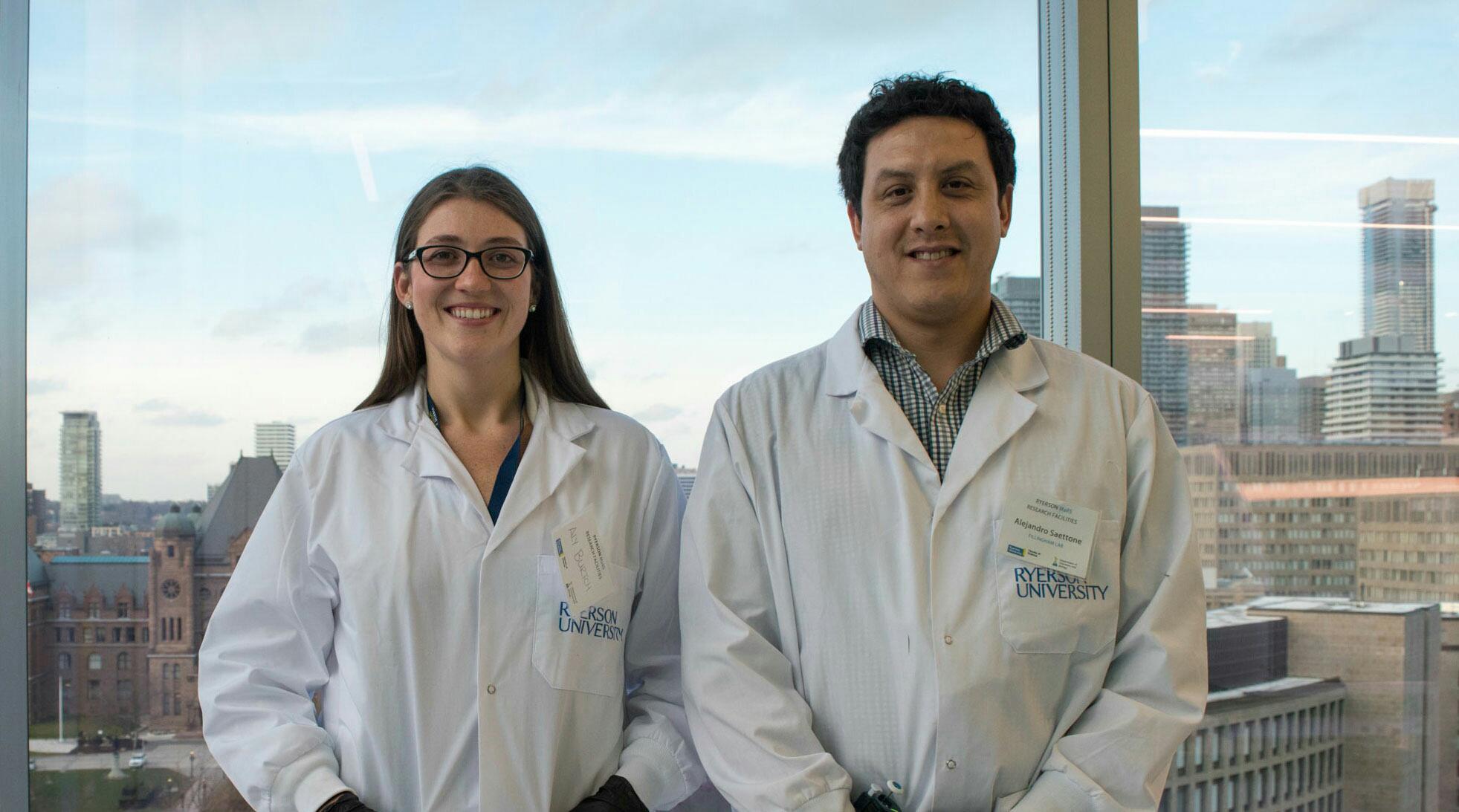Aly Burtch and Alejandro Saettone wear lab coats and stand side-by-side in front of a window.