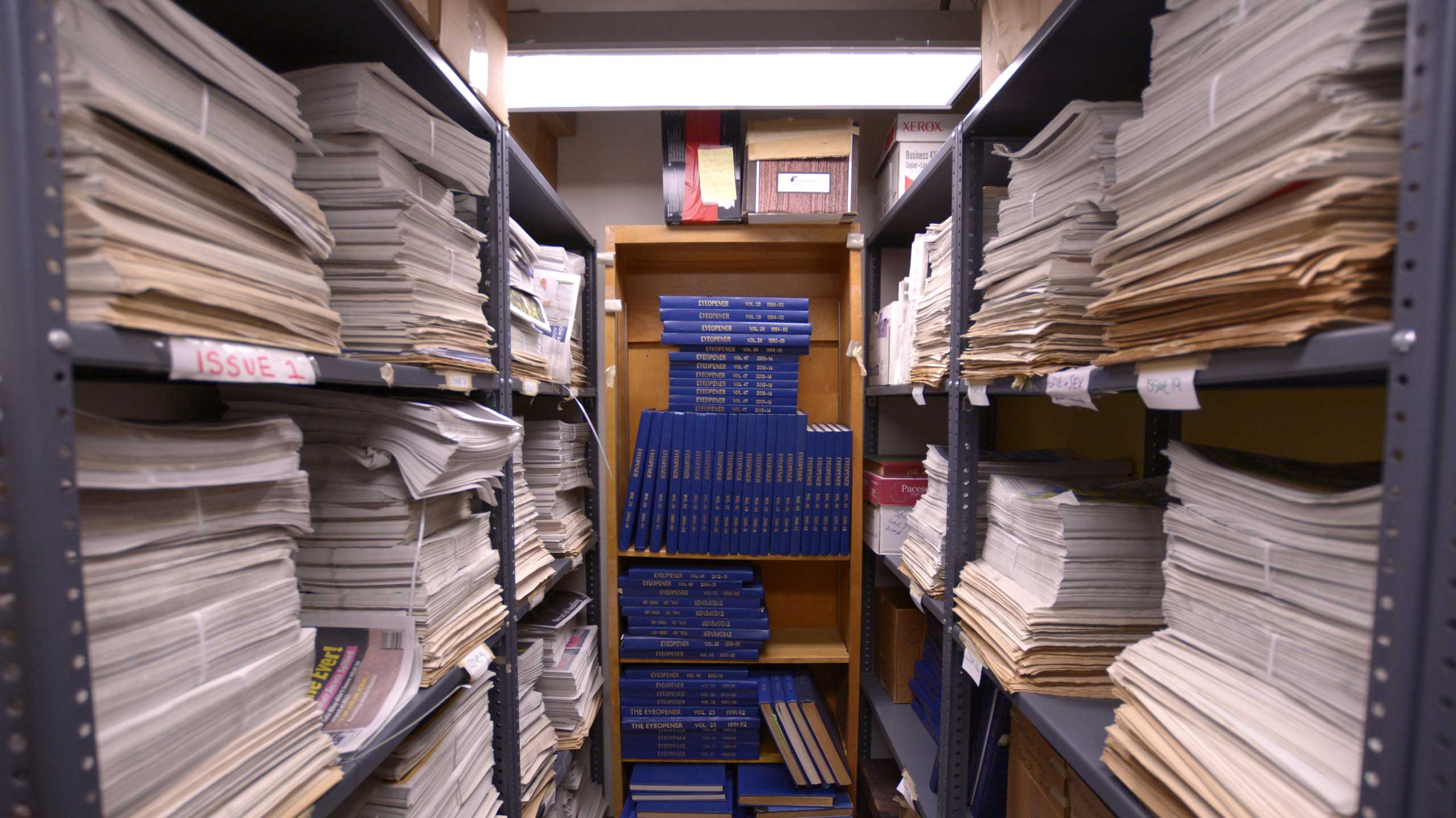 A look inside small room lined with floor to ceiling shelves, full of large stacks of newspapers. At the back of the room is a shelf with blue books labelled as "Eyeopener" with different dates. The room is poorly lit but cozy.