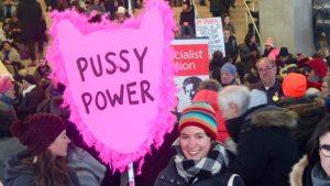 A woman stands in the Internation Women's Day crowd holding a pink fluffy sign that reads: Pussy Power