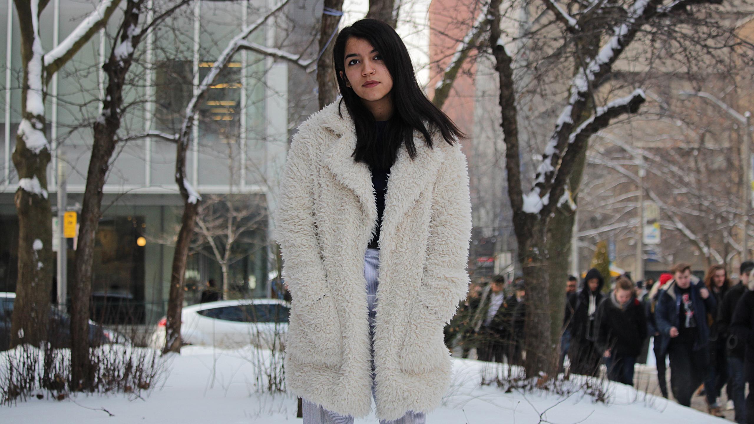 standing on Ryerson campus is a person wearing a fur coat. The background is a snowy winter wonderland.