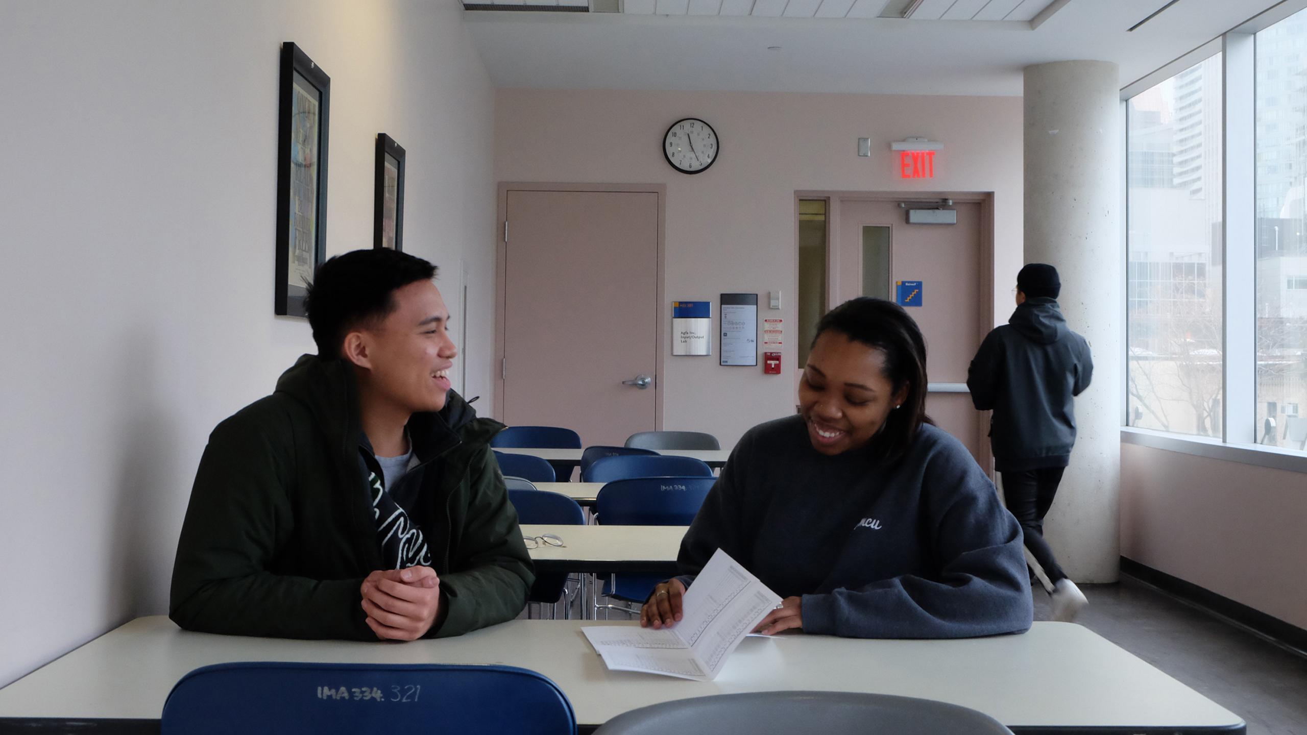 two students sitting at a table laughing and reading a pamphlet
