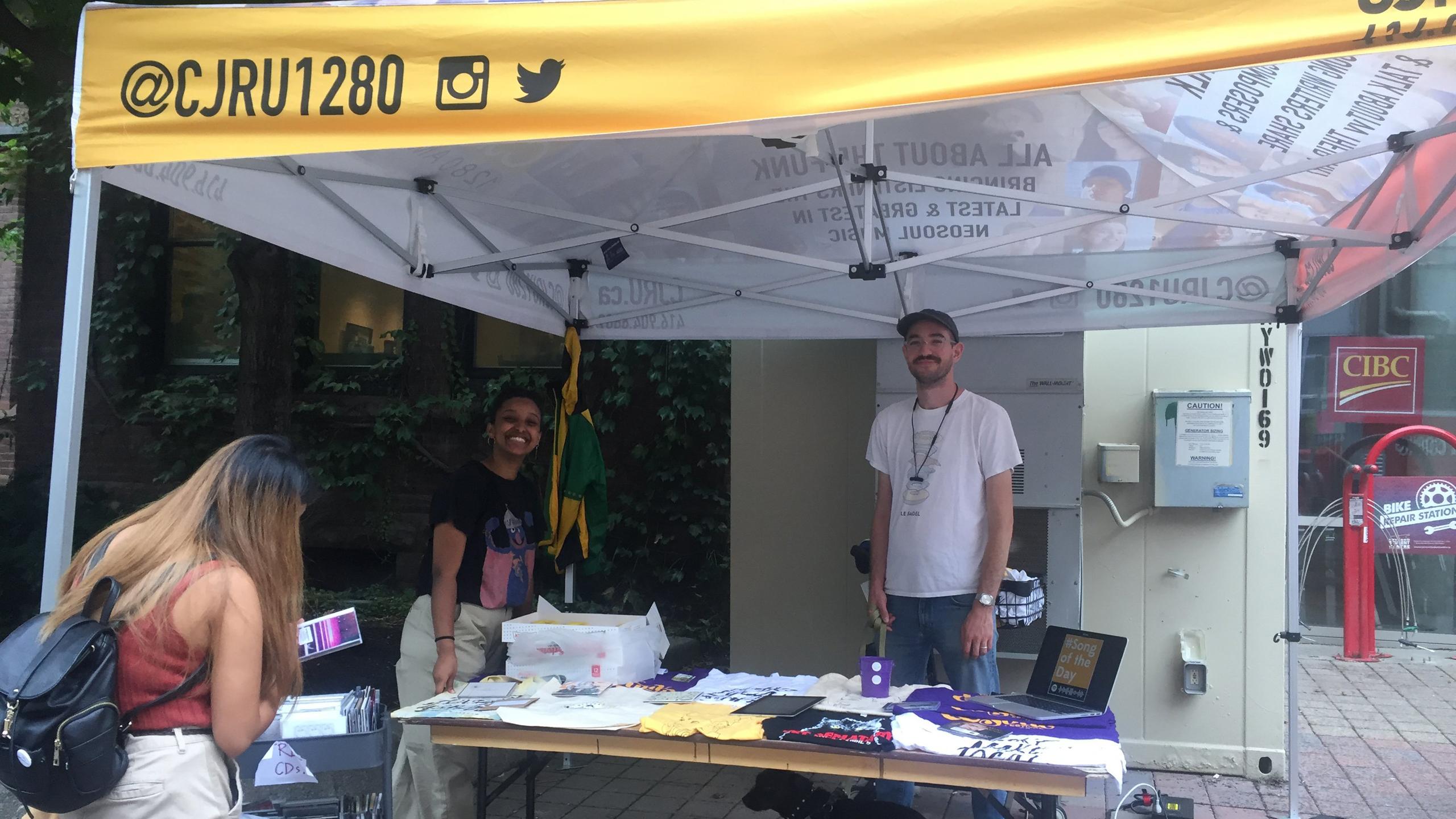 Two radio station workers stand behind a table of t-shirts and other free apparel underneath a yellow tent labelled CJRU.