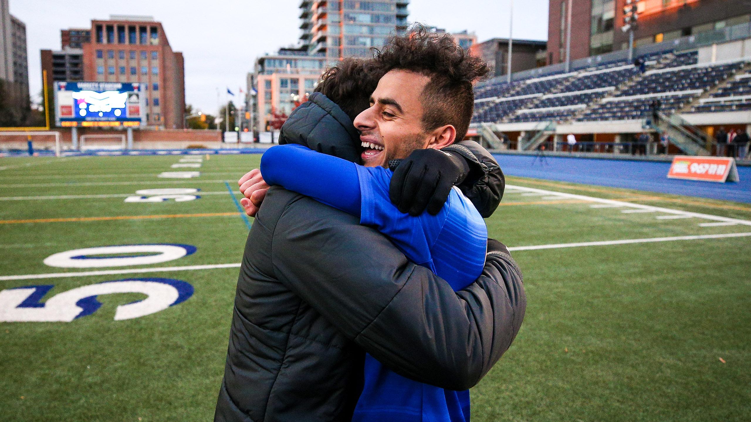 a man wearing a jacket is embraced by a soccer player wearing a blue jersey