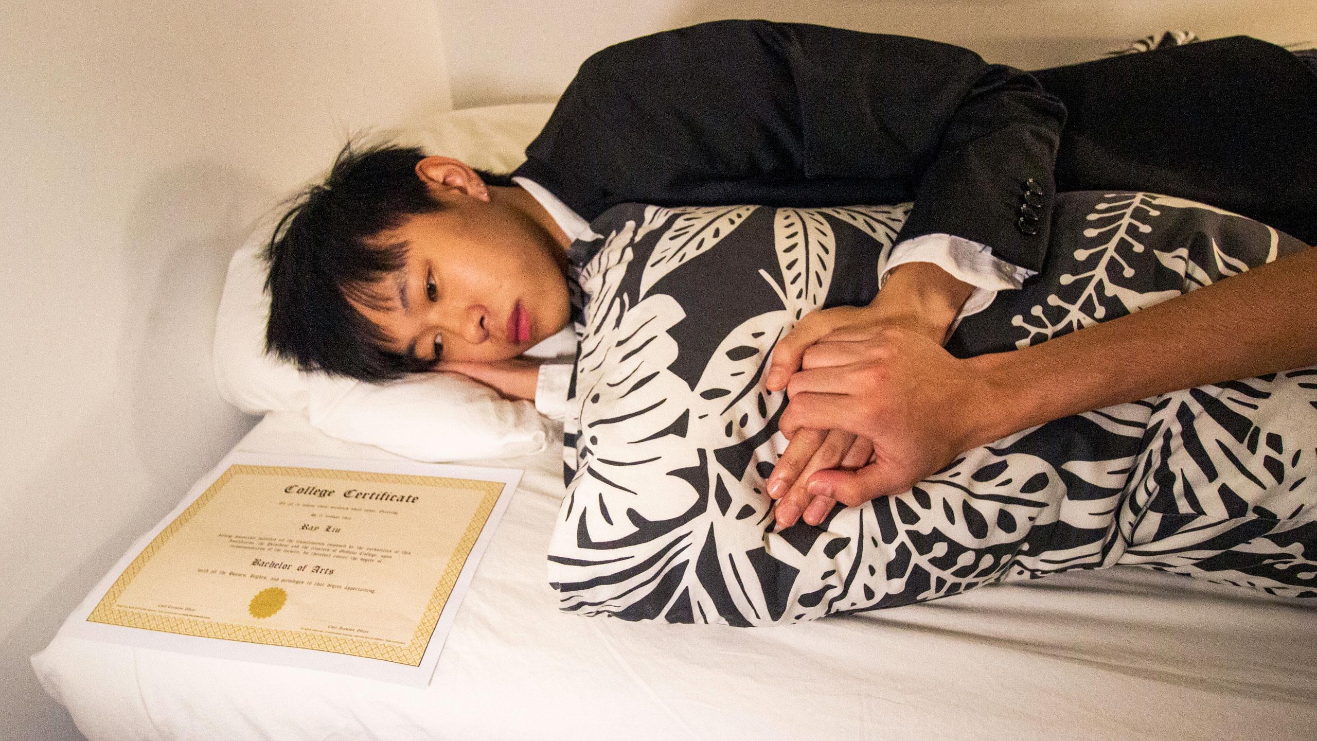 Student lies in bed solemnly with a degree next to him