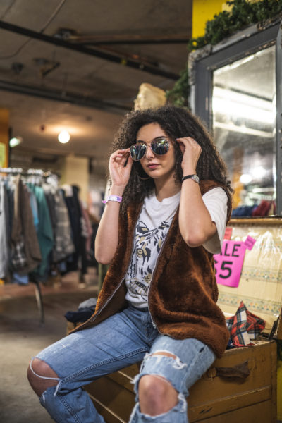 A person sits on a trunk of clothing in Black Market. They have sunglasses on their face and are wearing a fur vest, ripped jeans and t-shirt with a tiger's face on it. 