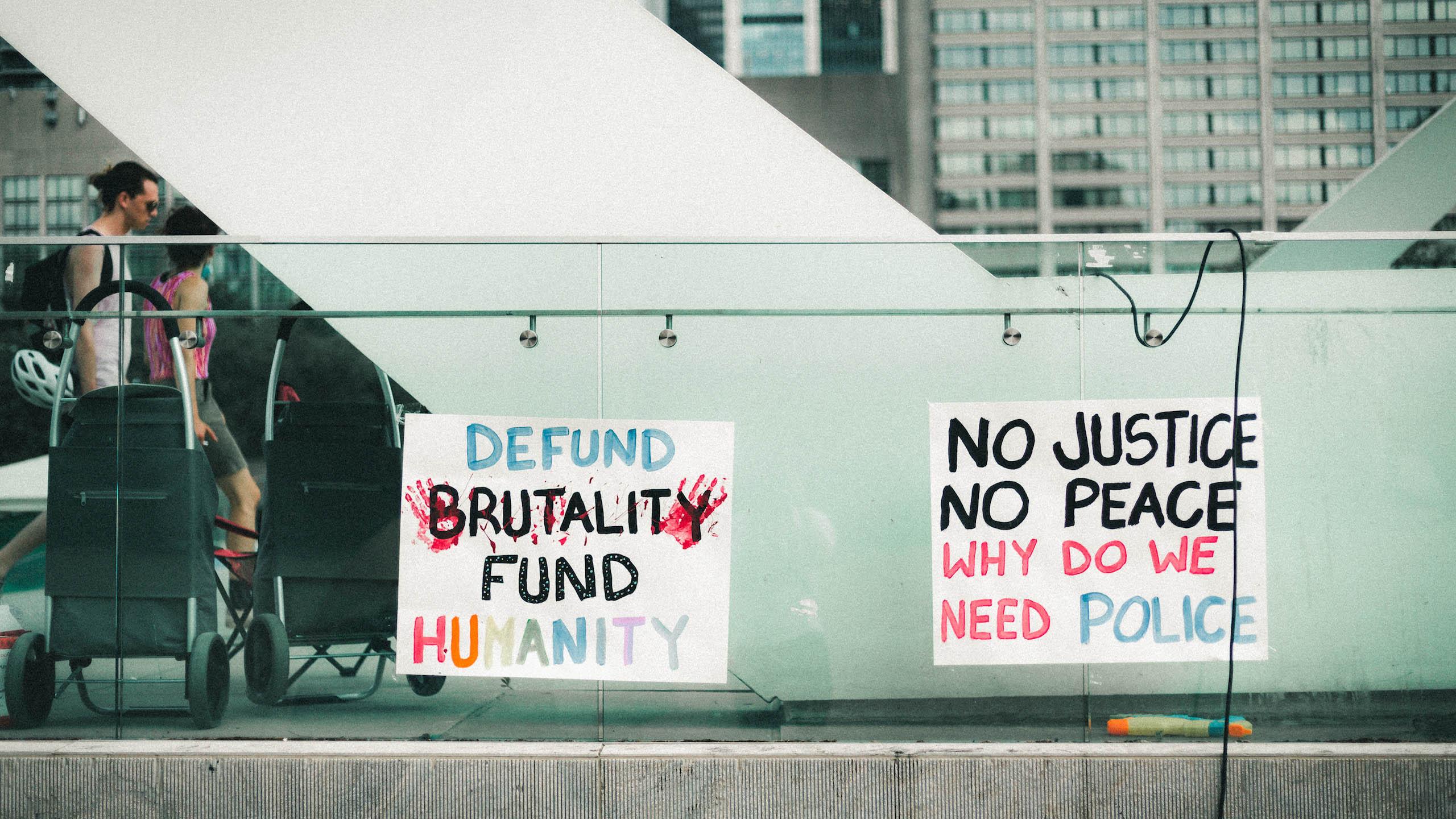 One sign reads: defund brutality, fund humanity; another sign reads no justice, no peace, why do we need police