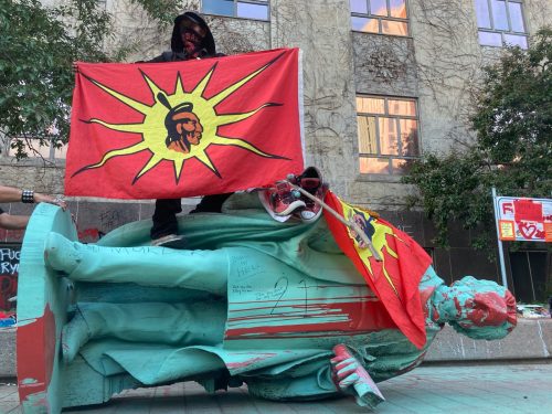 A protestor stands on top of the toppled Egerton Ryerson statue holding an Indigenous unity flag.