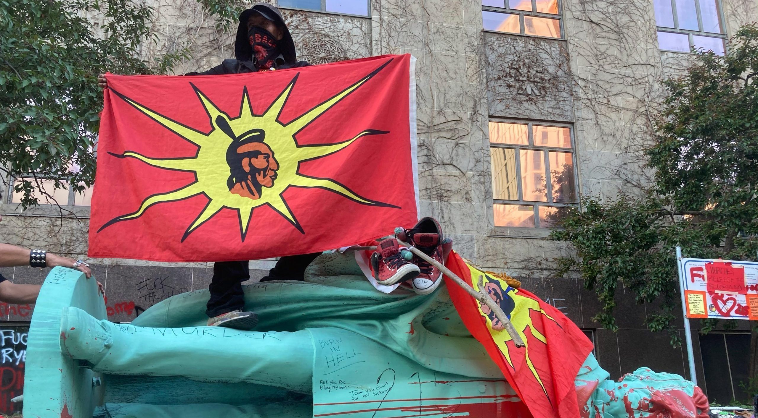 A protestor stands on top of the toppled Egerton Ryerson statue holding an Indigenous unity flag.