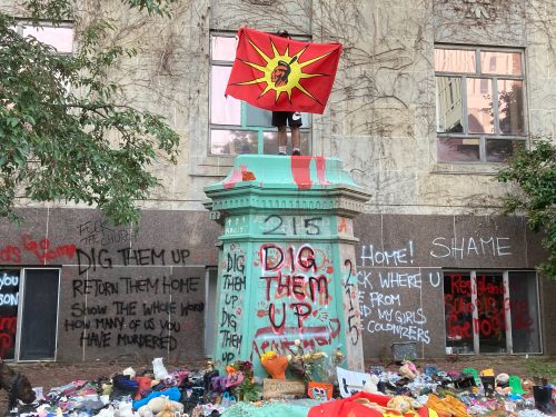 A protestor stands on the base of the Egerton Ryerson statue holding an Indigenous unity flag. The base is surrounded by flowers and pairs of children’s shoes. Writing on the wall behind the base it says, “dig them up, return them home, show the whole world how many of us you have murdered.”