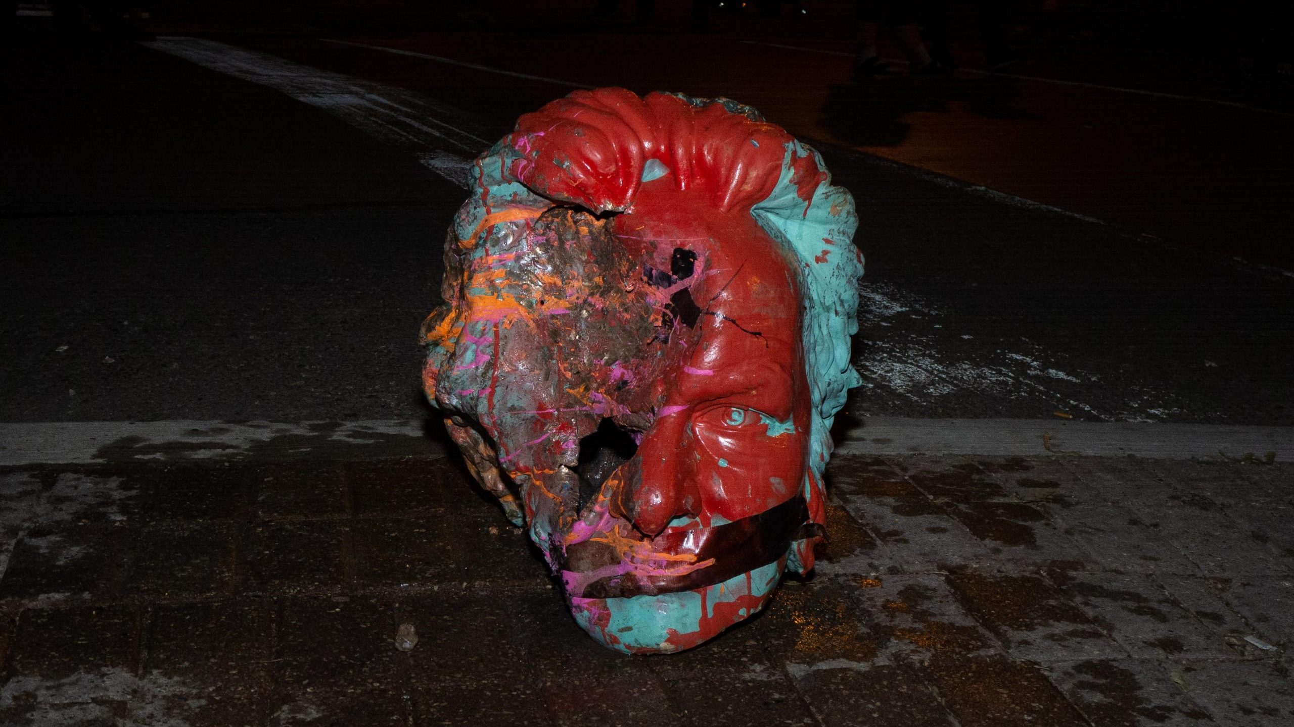 The decapitated head of the Egerton Ryerson statue sitting on the sidewalk with tape over its mouth. The side of the face is bashed in and the head is covered in paint.