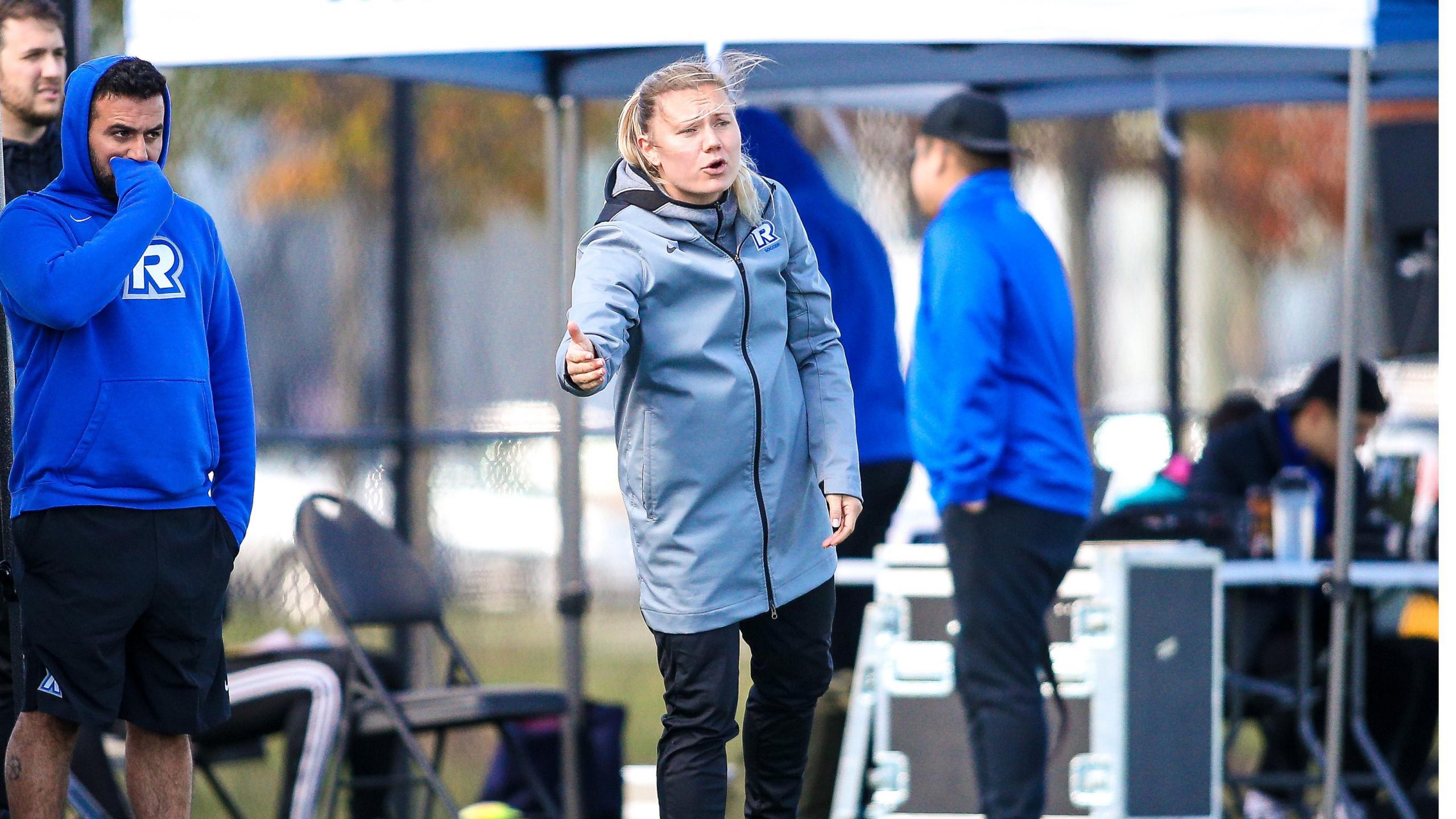 A woman in a grey jacket points to the field with a frustrated expression on her face