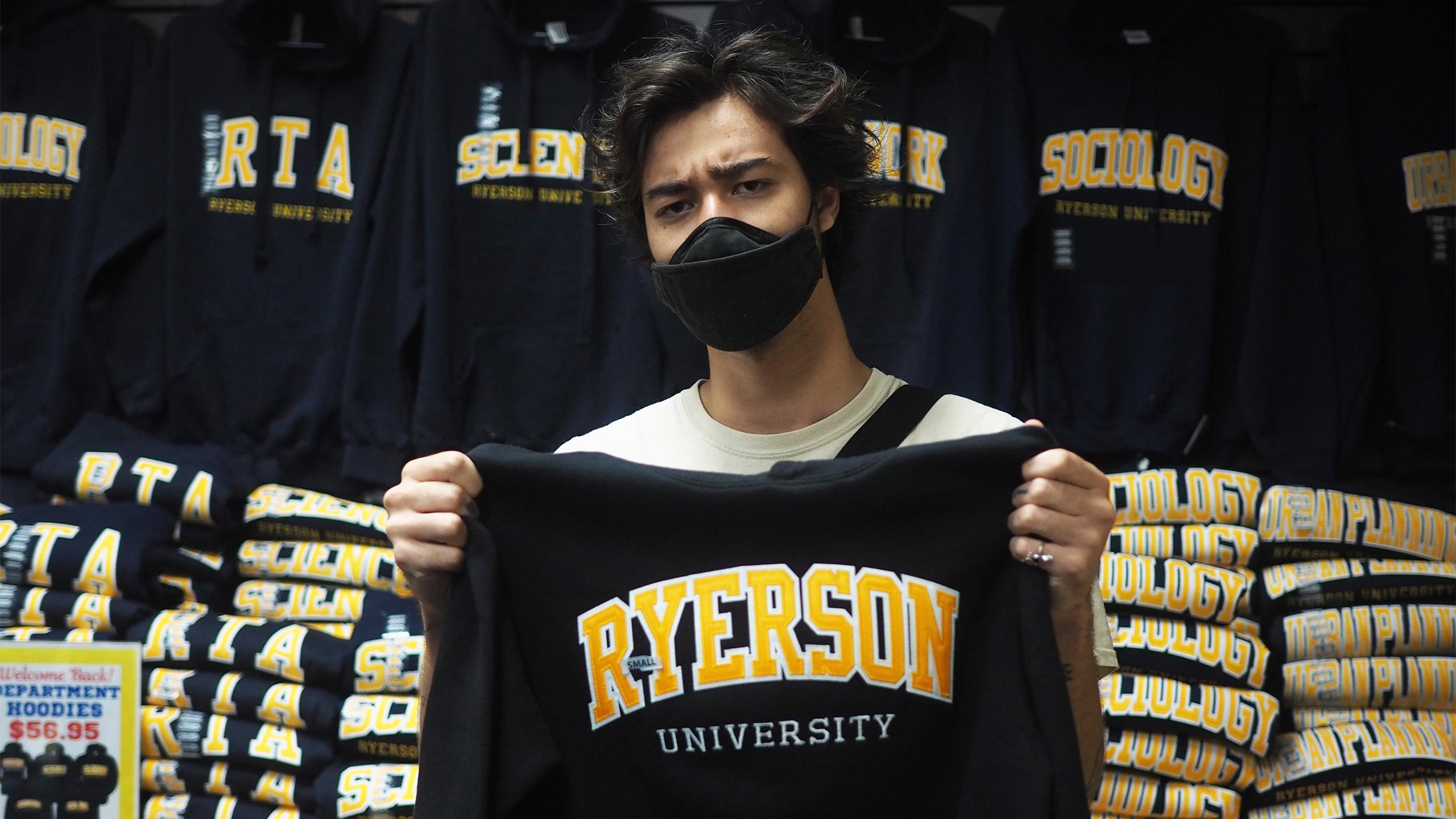 A man scowling holding up a Ryerson hoodie in the campus store