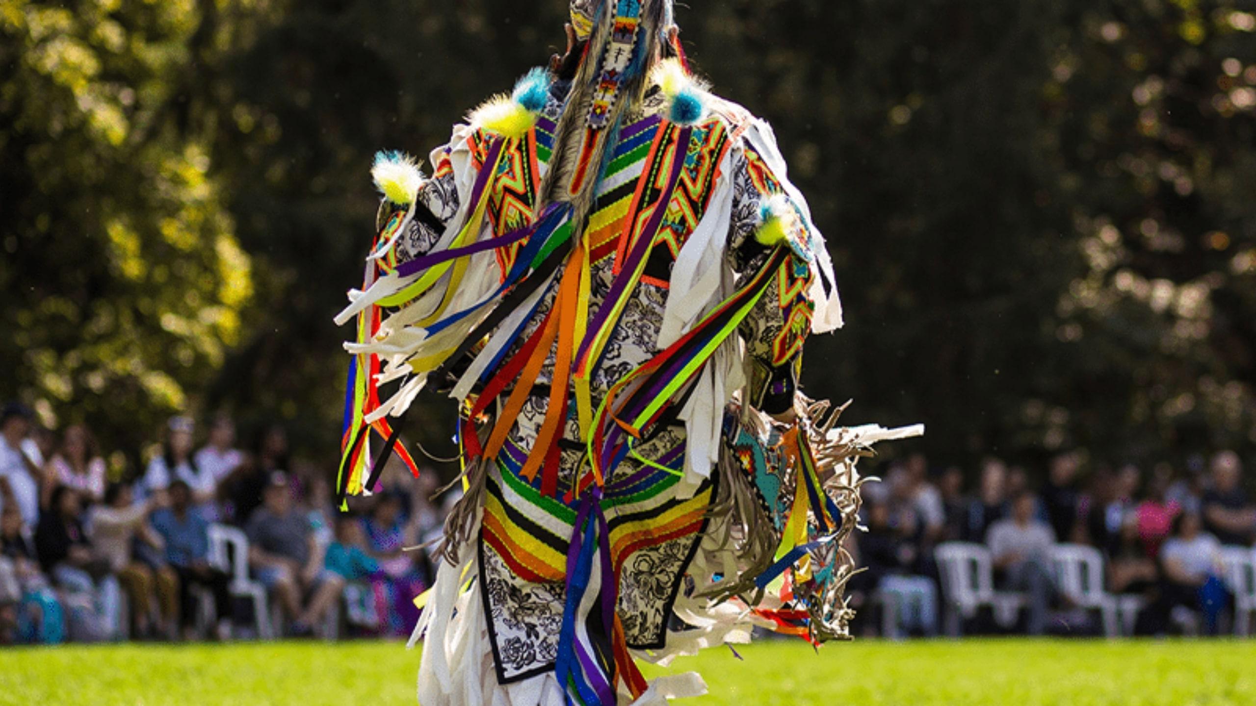An Indigenous man in a traditional outfit outside in a green space during a past, in-person pow wow