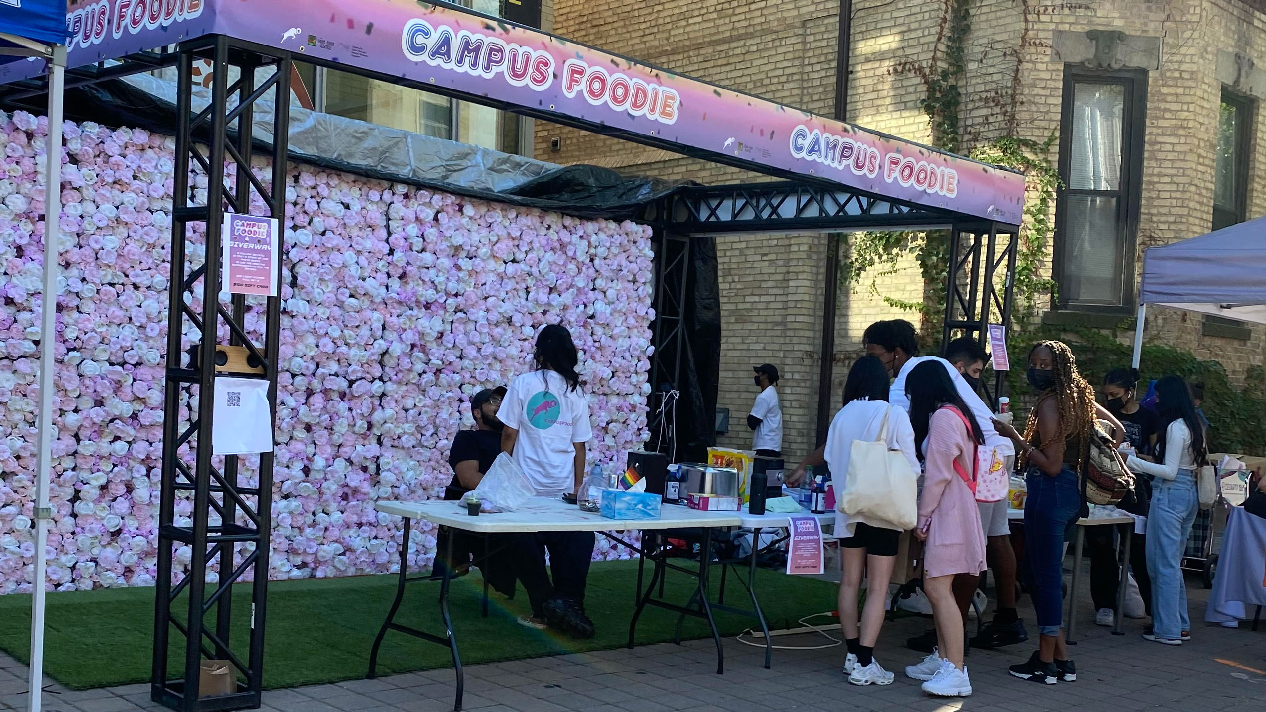 The Ryerson Students' Union table outside of the Ryerson Student Centre during Orientation Week in August 2021. Students are lined up for free food in front of the table while one person sits on another person's lap behind the table, in front of a large rose wall.