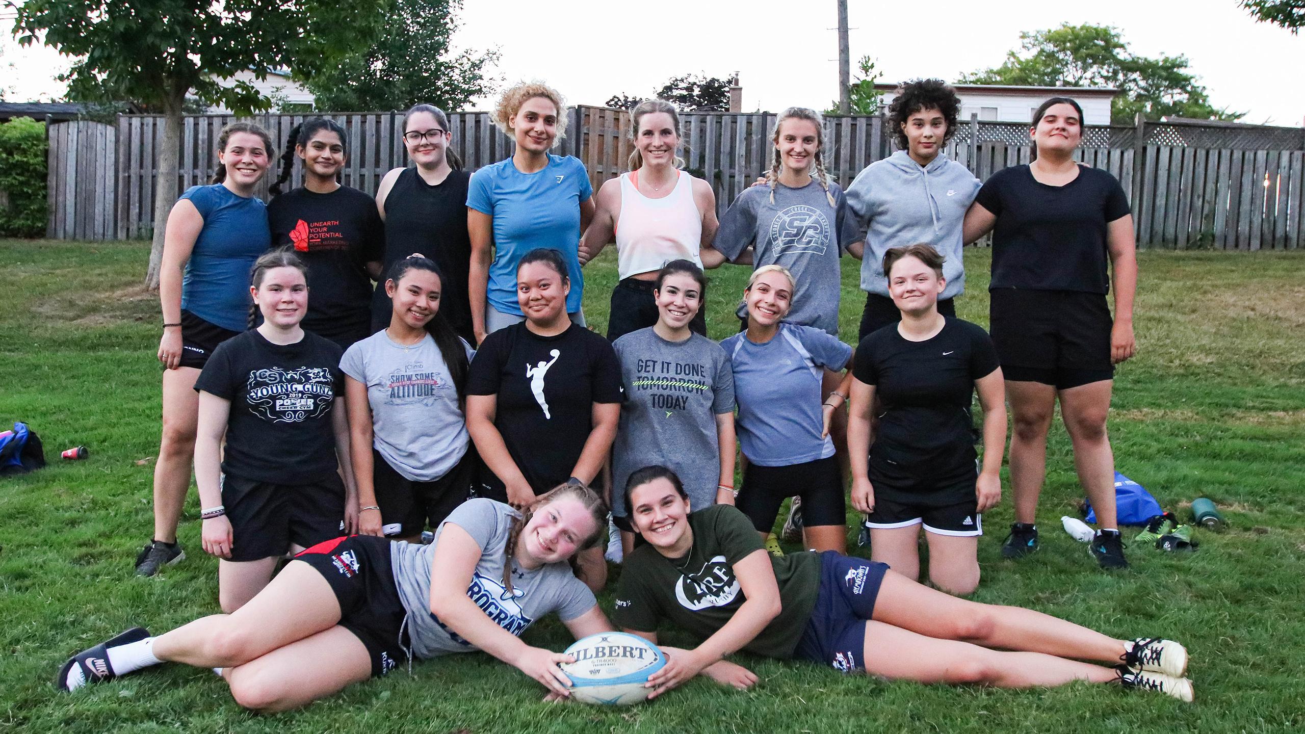 The Ryerson Rams women's rugby team, smiling and posing outside in the grass with a brown fence behind them. The back row of people are standing, the middle row are on their knees, and the front few are lying down, holding a rugby ball with the word "Gilbert" on it. The team consists of women from various ethnic backgrounds, but they're all young and in athletic wear.