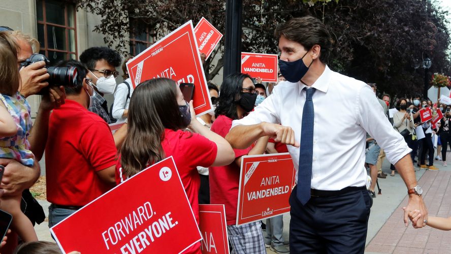Justin Trudeau in a black mask, white shirt, blue tie and black pants bumping elbows with supporters dressed in red Liberal party merch at a rally. Some attendees are holding signs that say "Forward for everyone" or "Anita Vandenbeld"