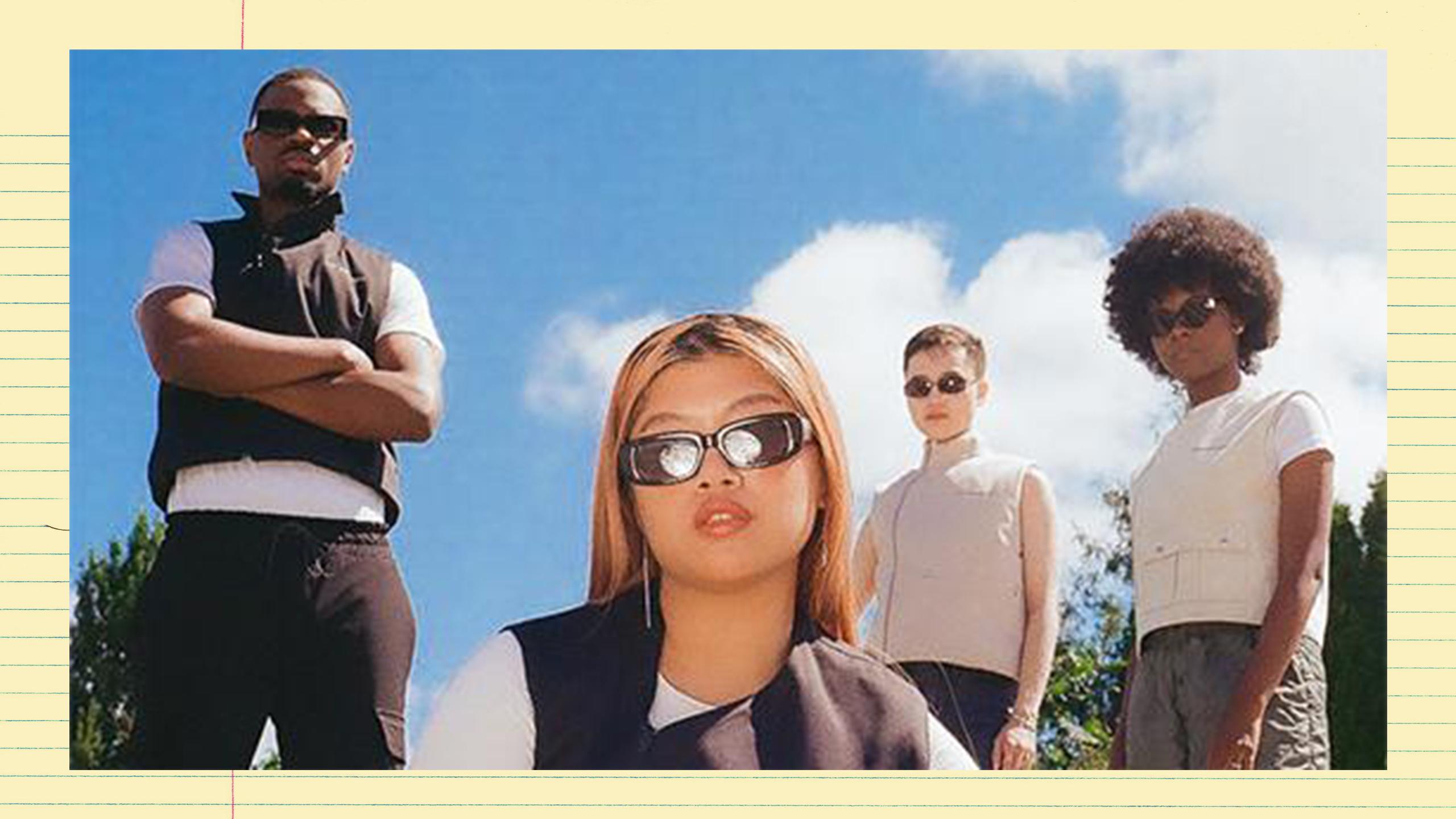 A group of people wearing sunglasses and black, white and cream athleisure, looking at the camera. They're standing outside with green trees and a blue, sunny sky behind them. The photo is taken from a low angle.