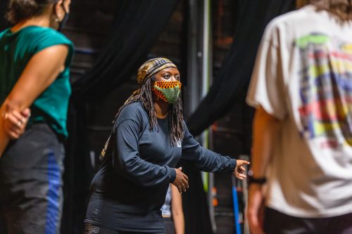 A woman wearing patterned mask and all black clothing leads a dance class