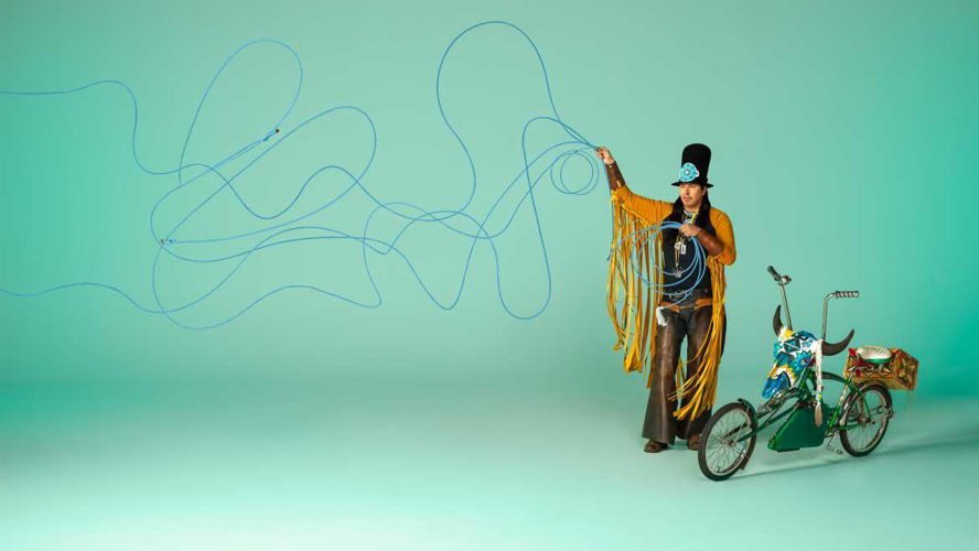 A man in Indigenous regalia stands next to a bicycle against a blue background