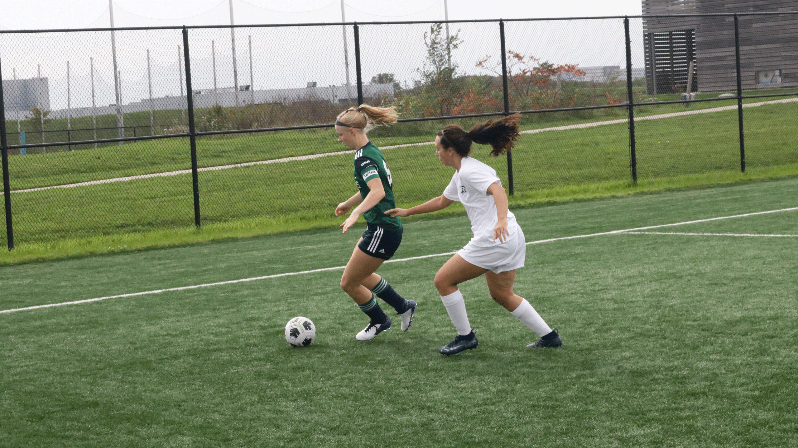 A soccer player with a white jersey fights for the ball with a soccer player in a green jersey