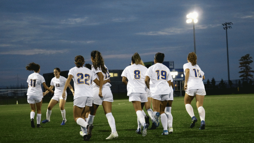 A group of soccer players wearing white jerseys come together to celebrate a goal