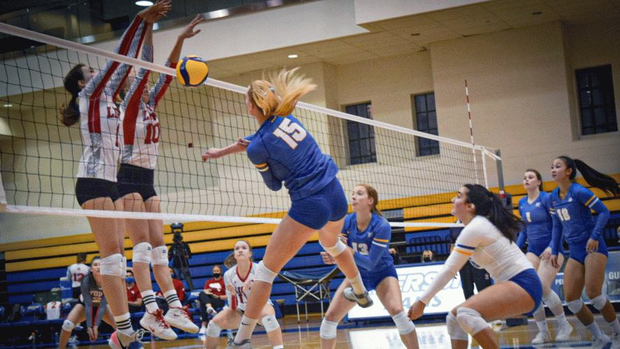 A Rams women's volleyball player in a blue jersey is blocked at the net