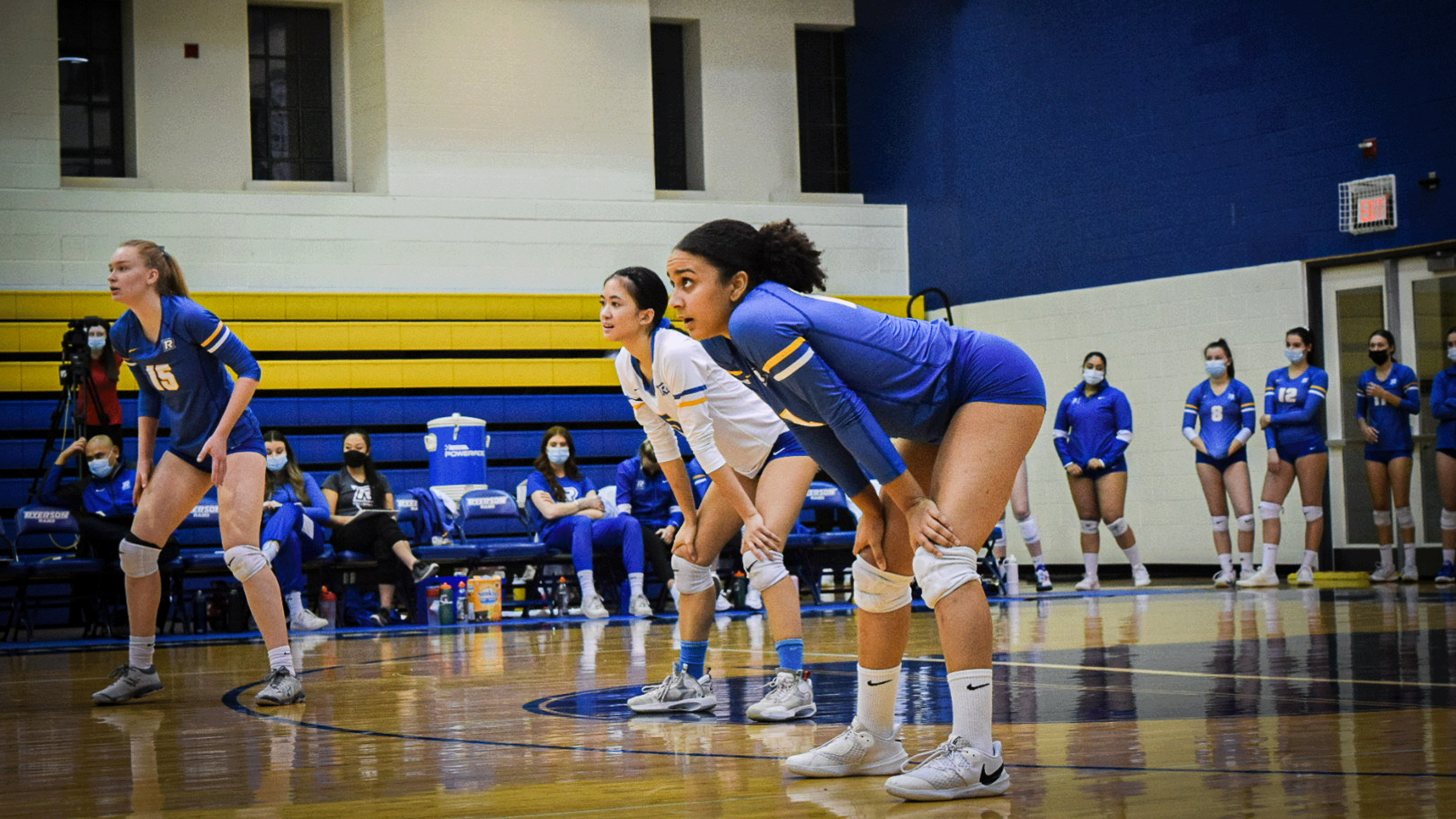 The Rams women's volleyball team in blue and white jerseys stand to receive a serve
