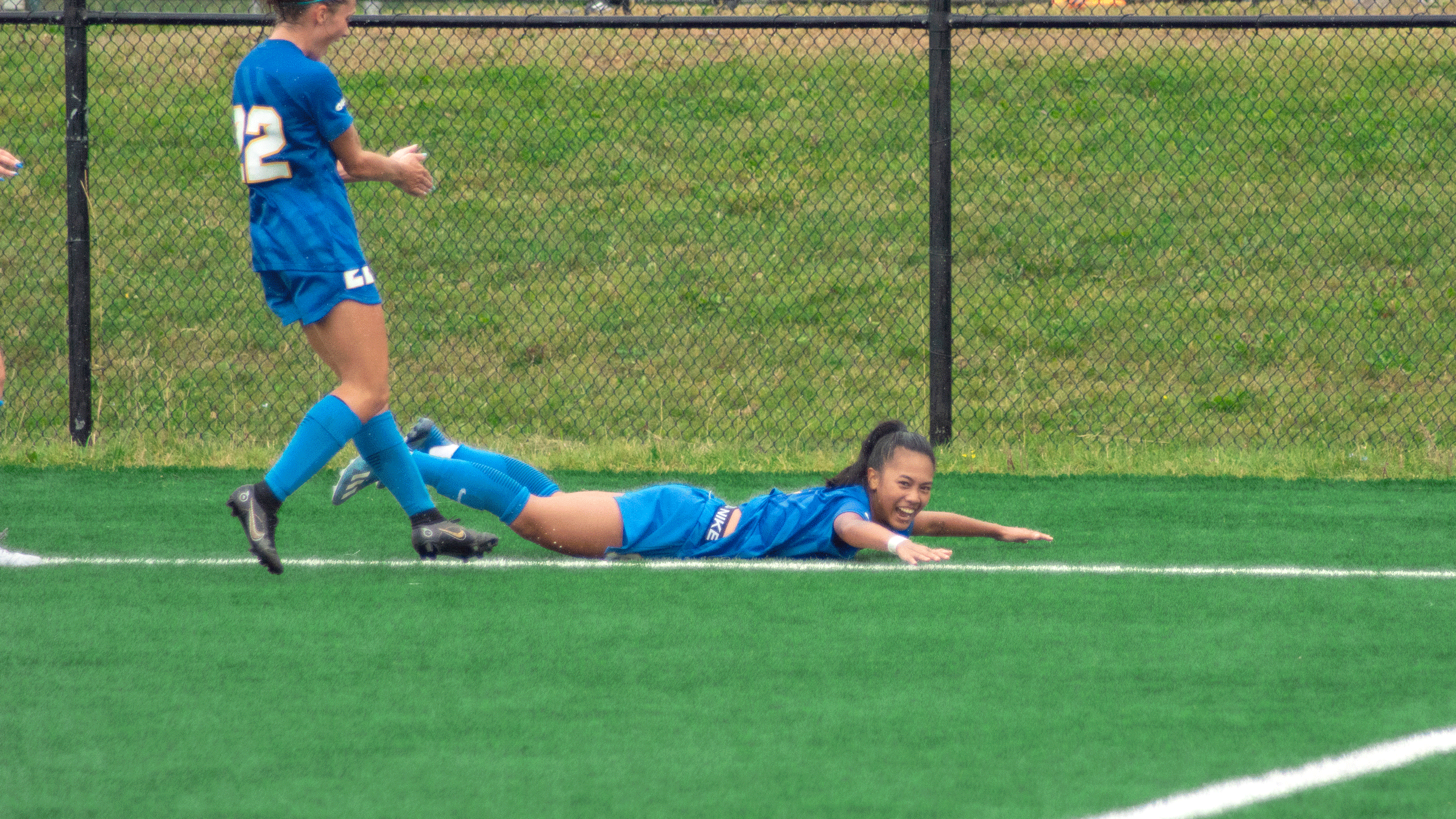 A soccer player in a blue jersey slides across the pitch with a smile on their face