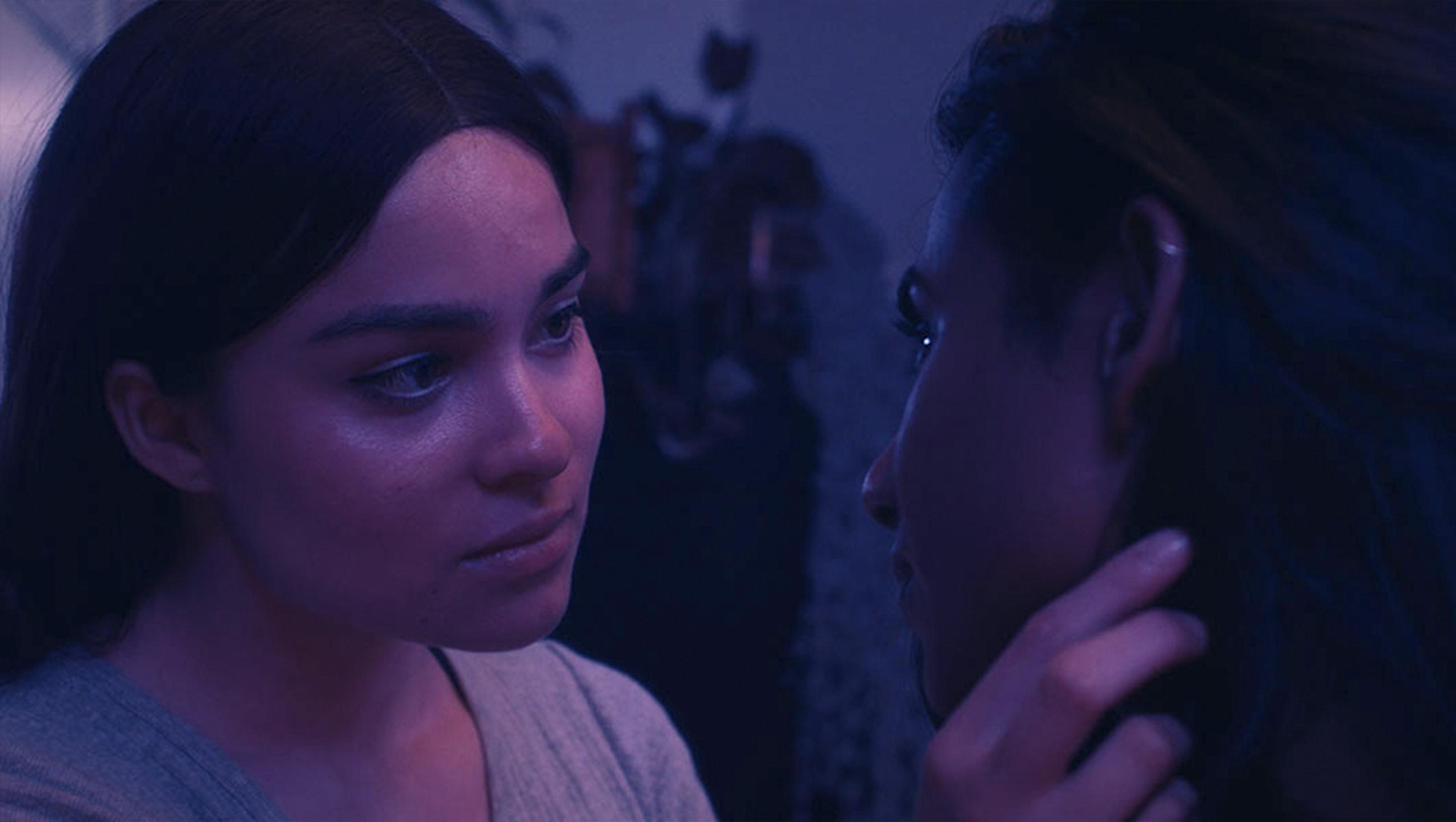 In a dark purple and pink lit room a woman to the left is tucking hair behind another woman’s ear on the right