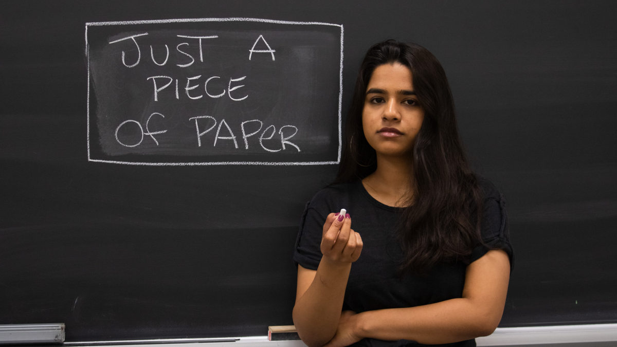 A student with no expression holding chalk in front of a chalkboard that reads “just a piece of paper”