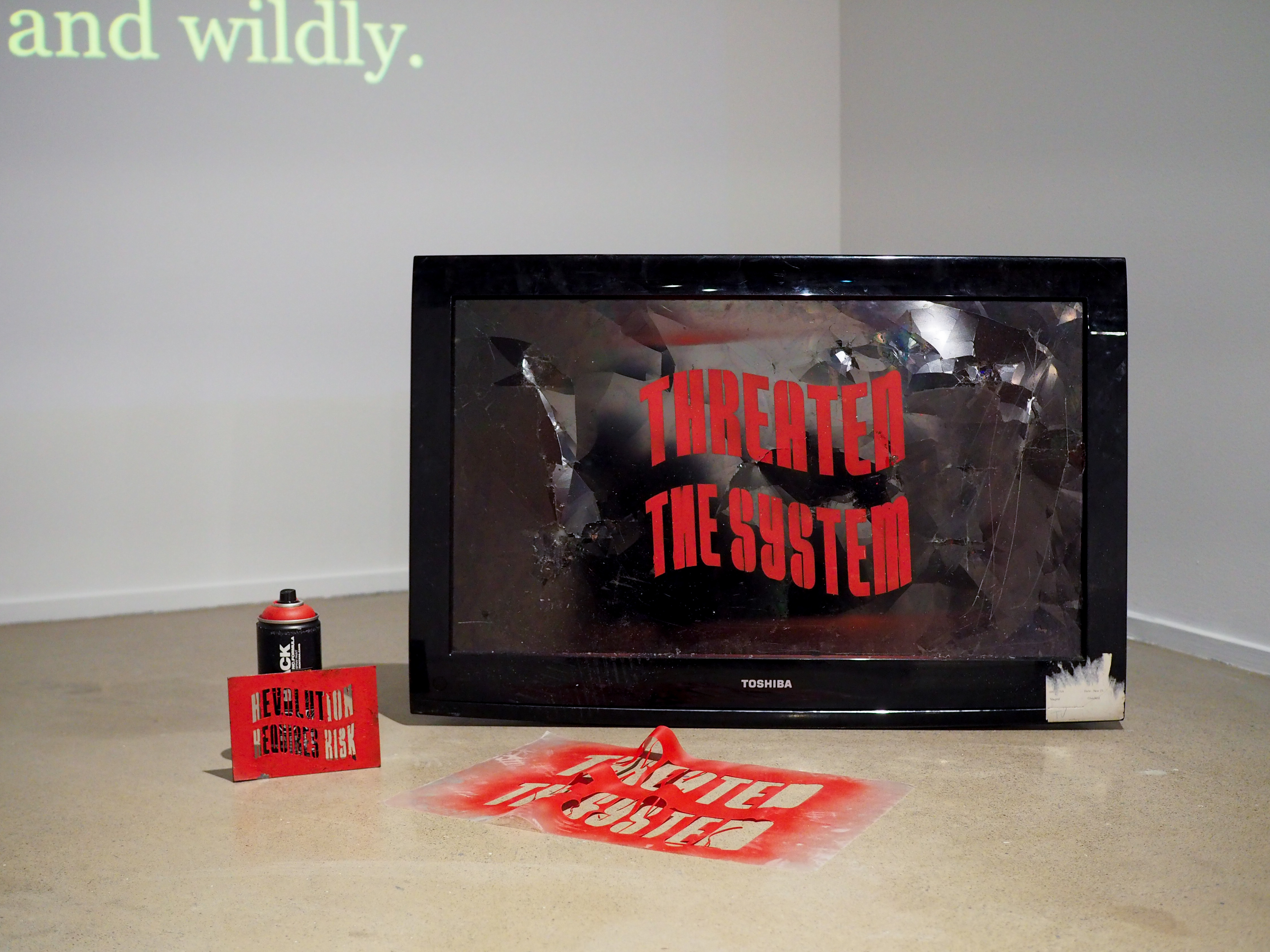 A broken television spray painted with a stencil that reads "THREATEN THE SYSTEM." Beside it, there's two used stencils and a can of spray paint.