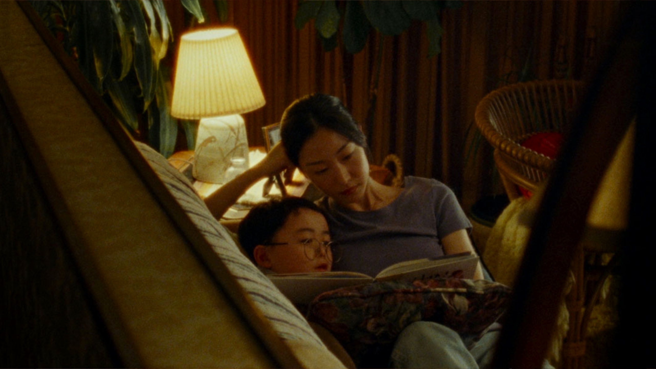 A mother and son laying on a couch reading a book in a dimly lit room