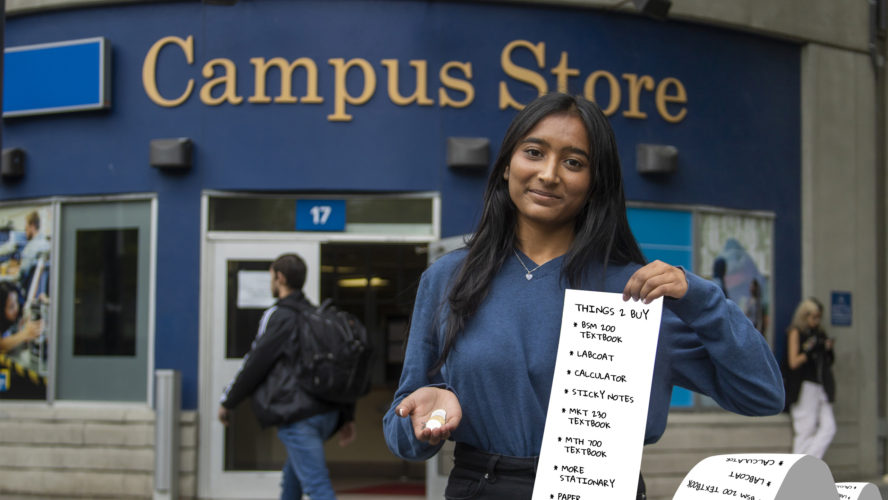 a student standing out front of the campus store, in one hand theres a handful of coins and in the other is a very long list of thigns to buy for school