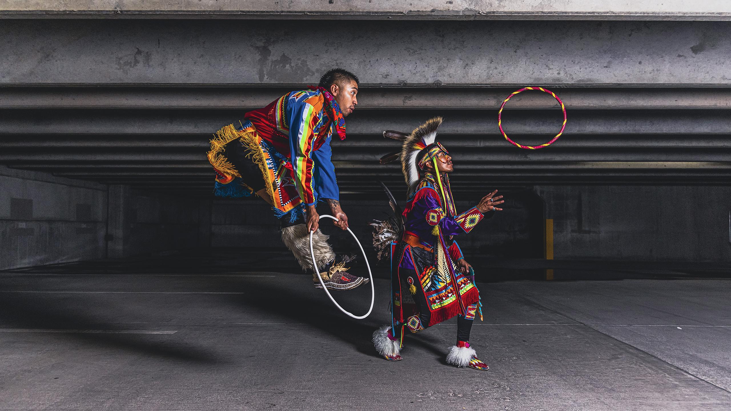 Two dancers of Indigenous Enterprise dancing in a parking garage with hoops. One dancer is jumping while the other is standing. 
