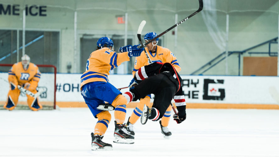 A TMU hockey player in a yellow jersey finishes a bodycheck on a UNB forward