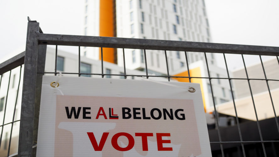 A sign hung on a fence that says "We all belong" and the word "Vote" below.