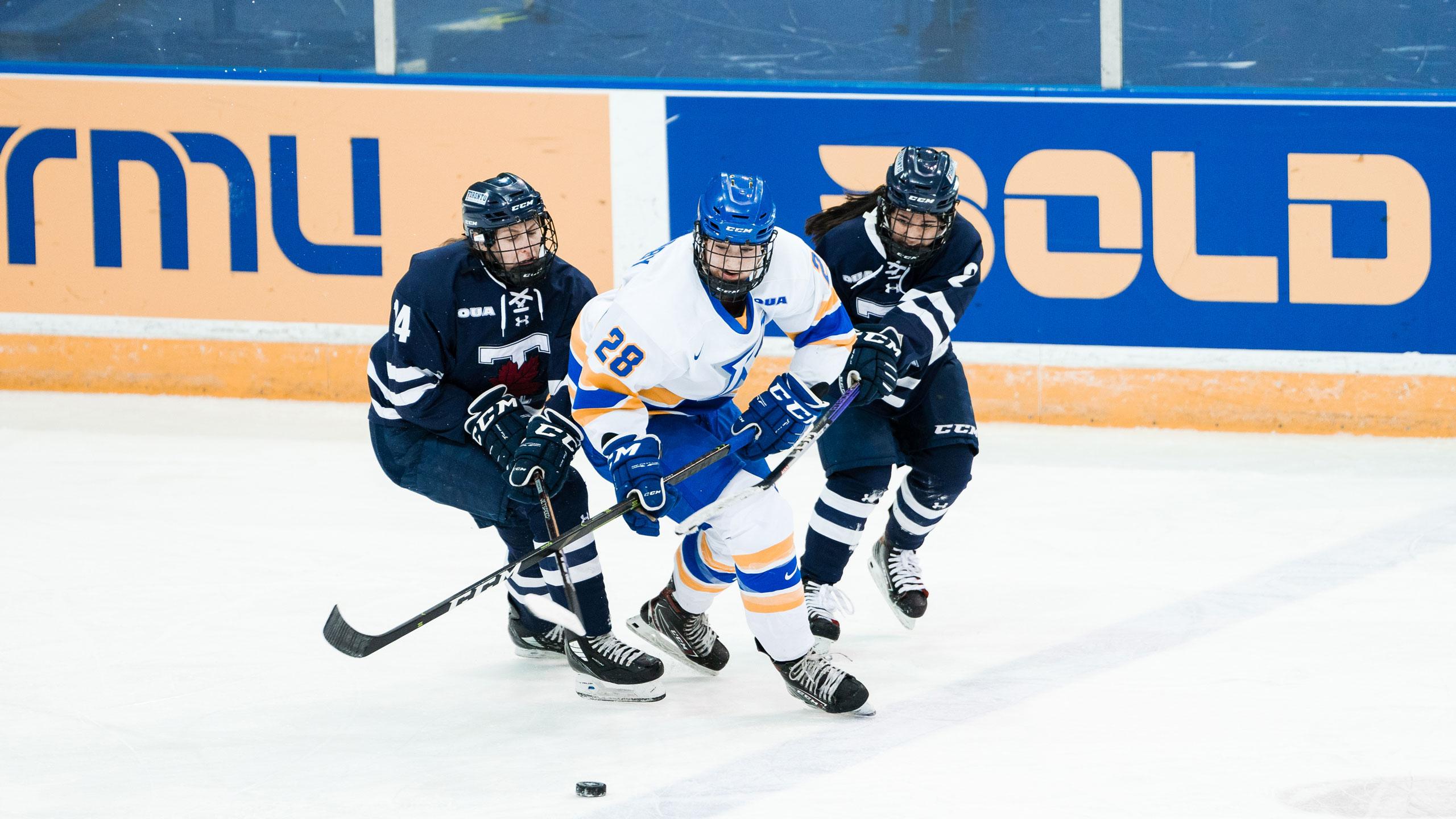 A TMU women's hockey player in a white jersey skates with the puck in front of two U of T players in dark blue jerseys