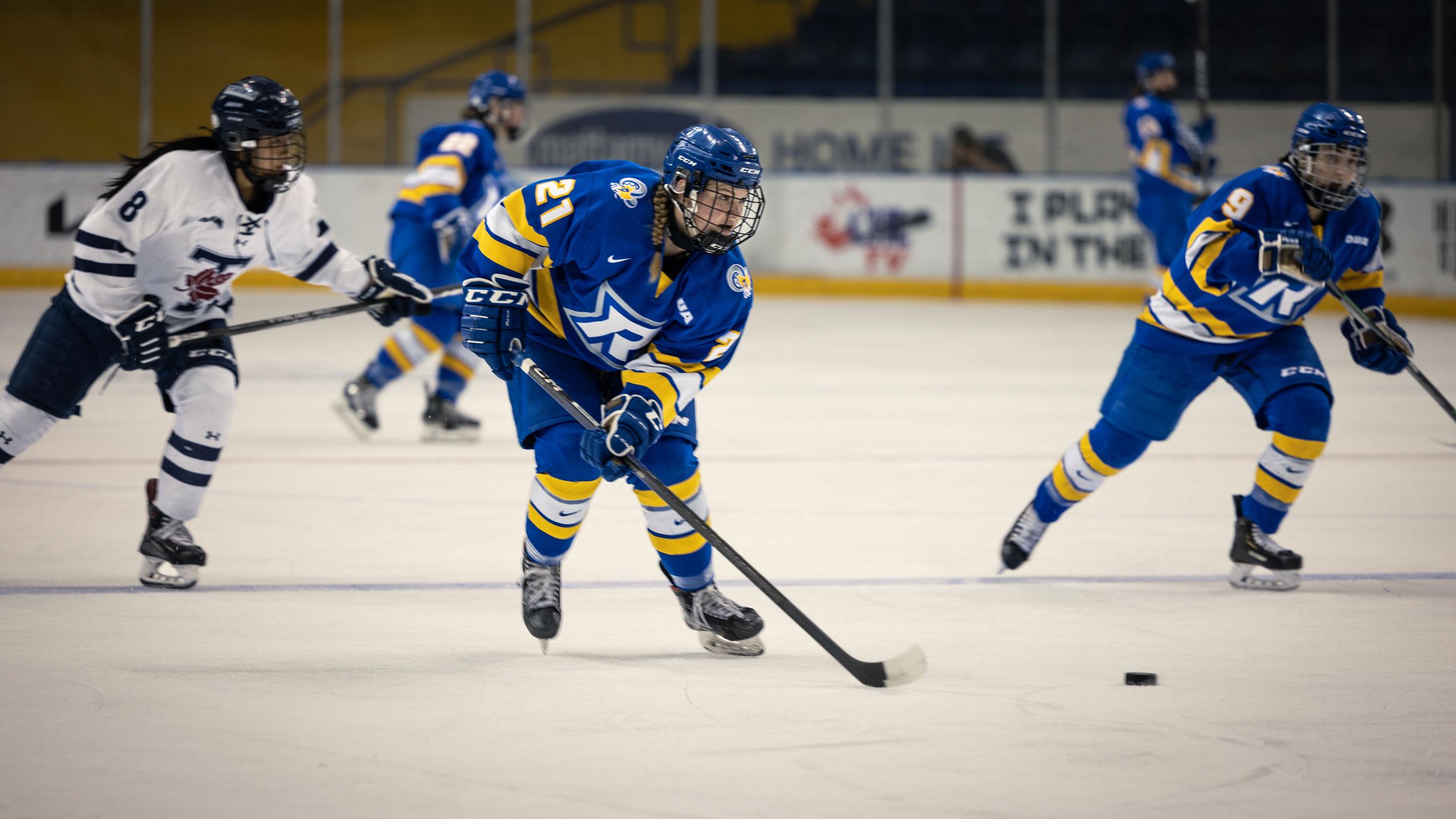 A TMU Bold women's hockey player in a blue jersey skates up the ice with the puck