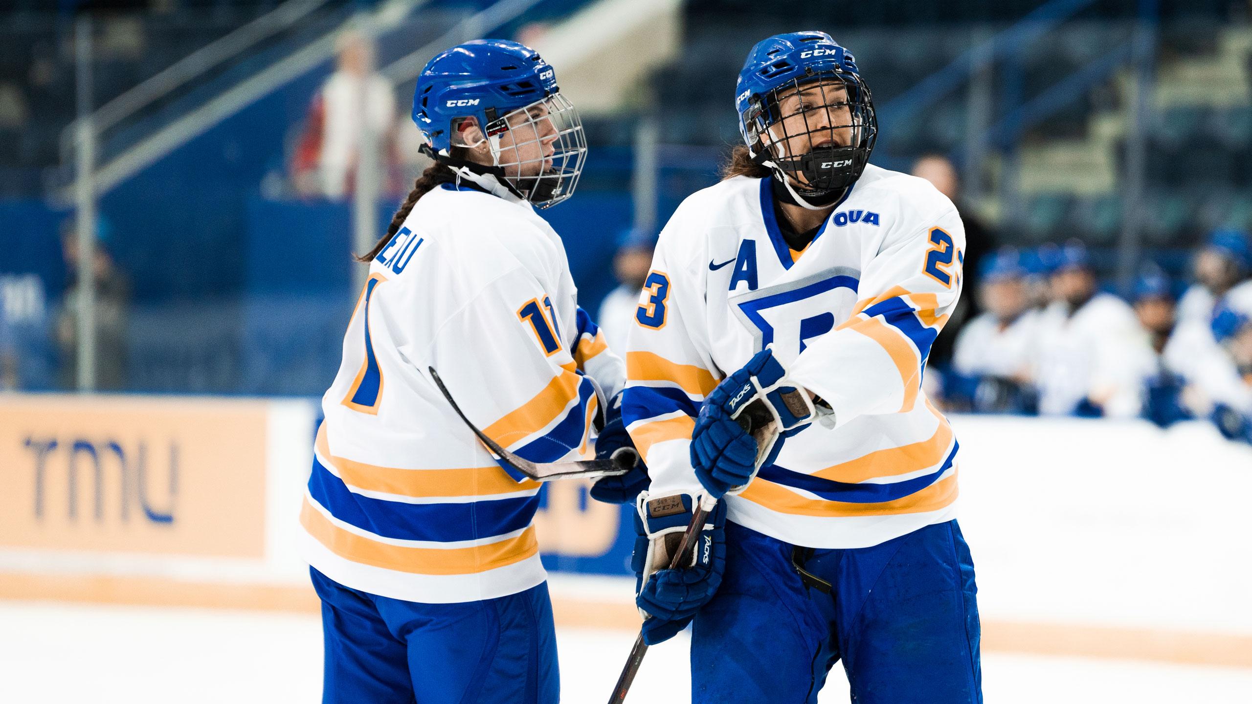 Two TMU women's hockey players in white jerseys talk to one another near centre ice