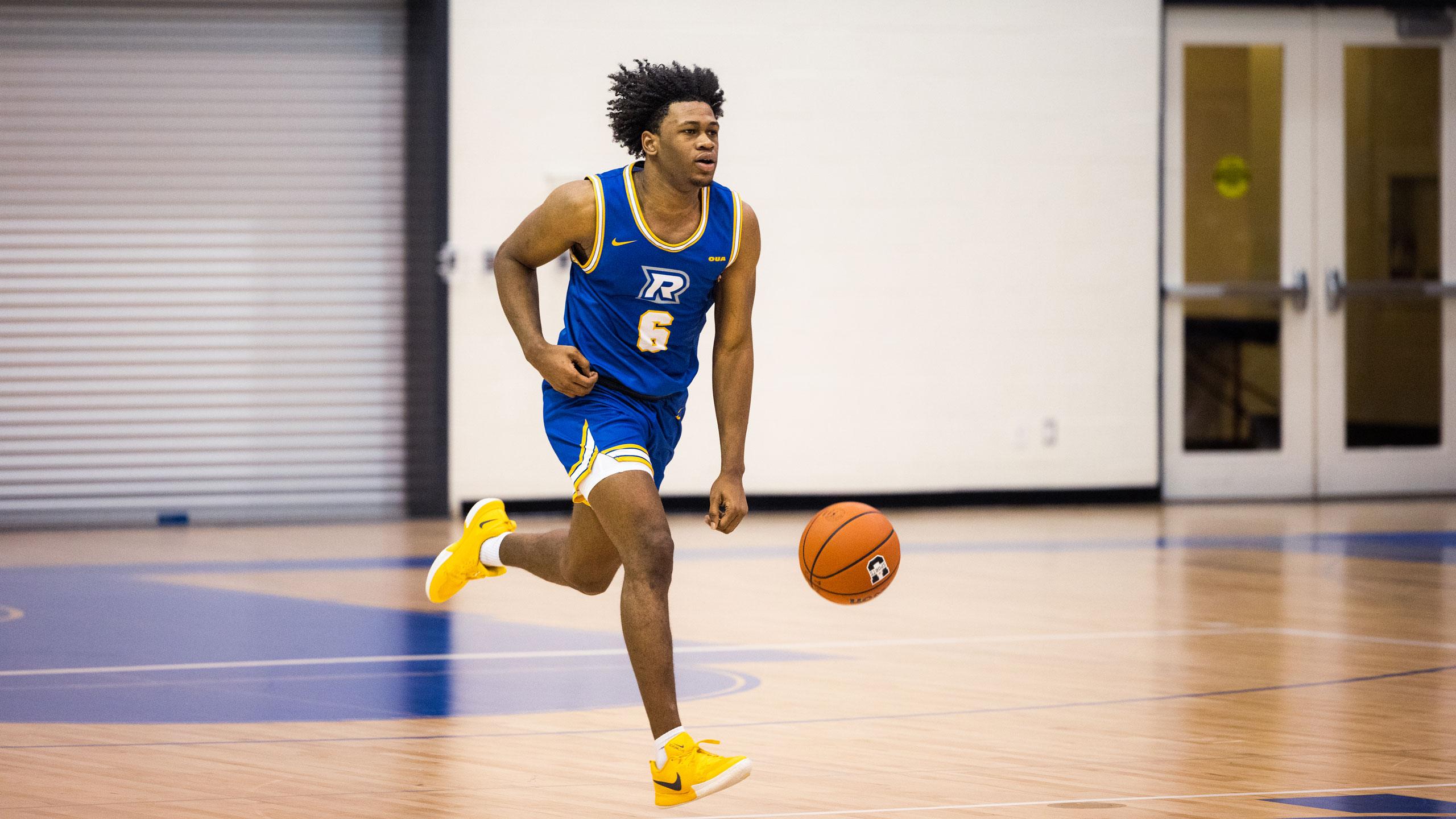 A TMU men's basketball player in a blue jersey dribbles the ball up the court