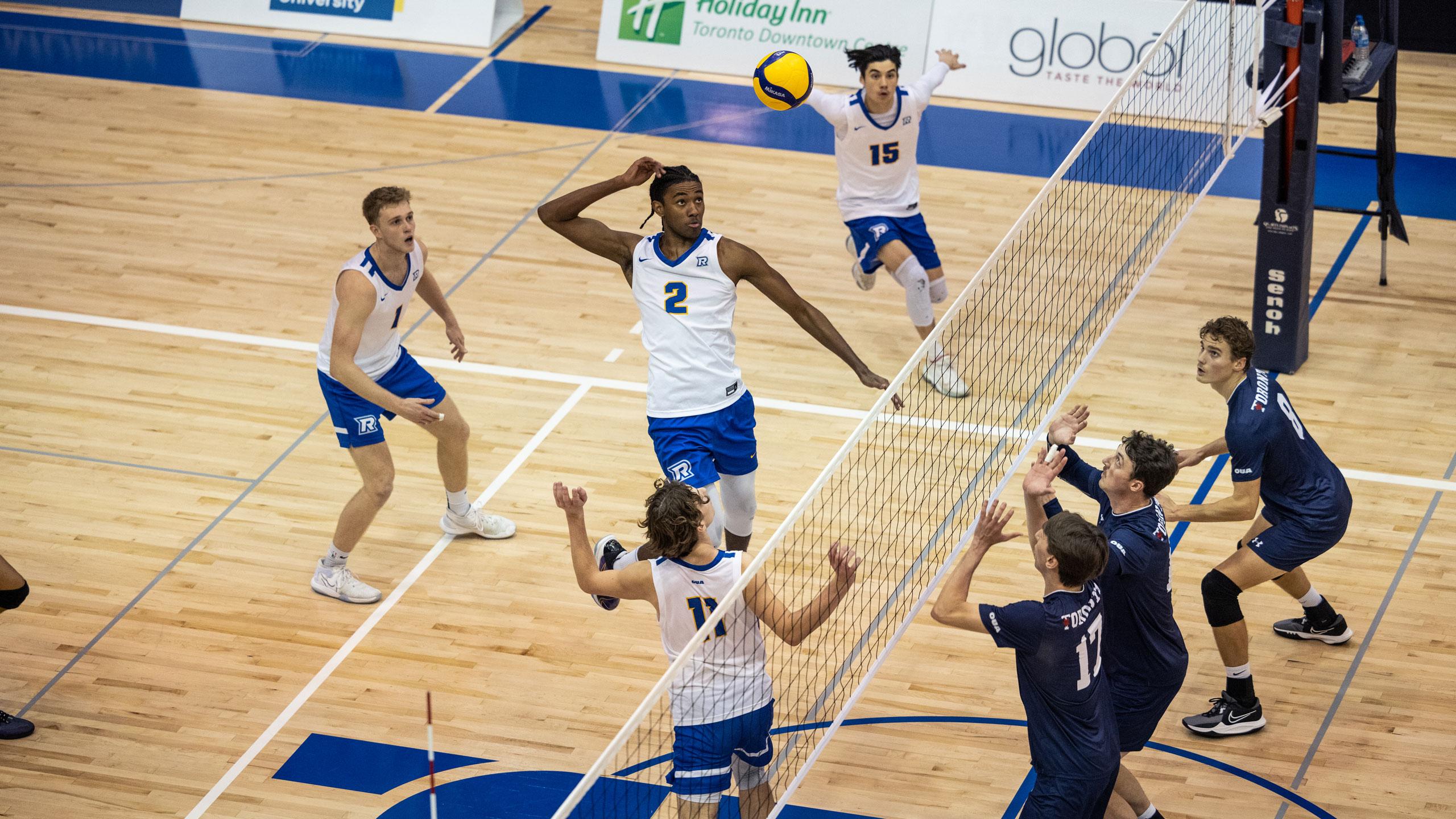 Four TMU men's volleyball players in white jerseys spike a ball over the net with three U of T players with dark blue jerseys on the other side