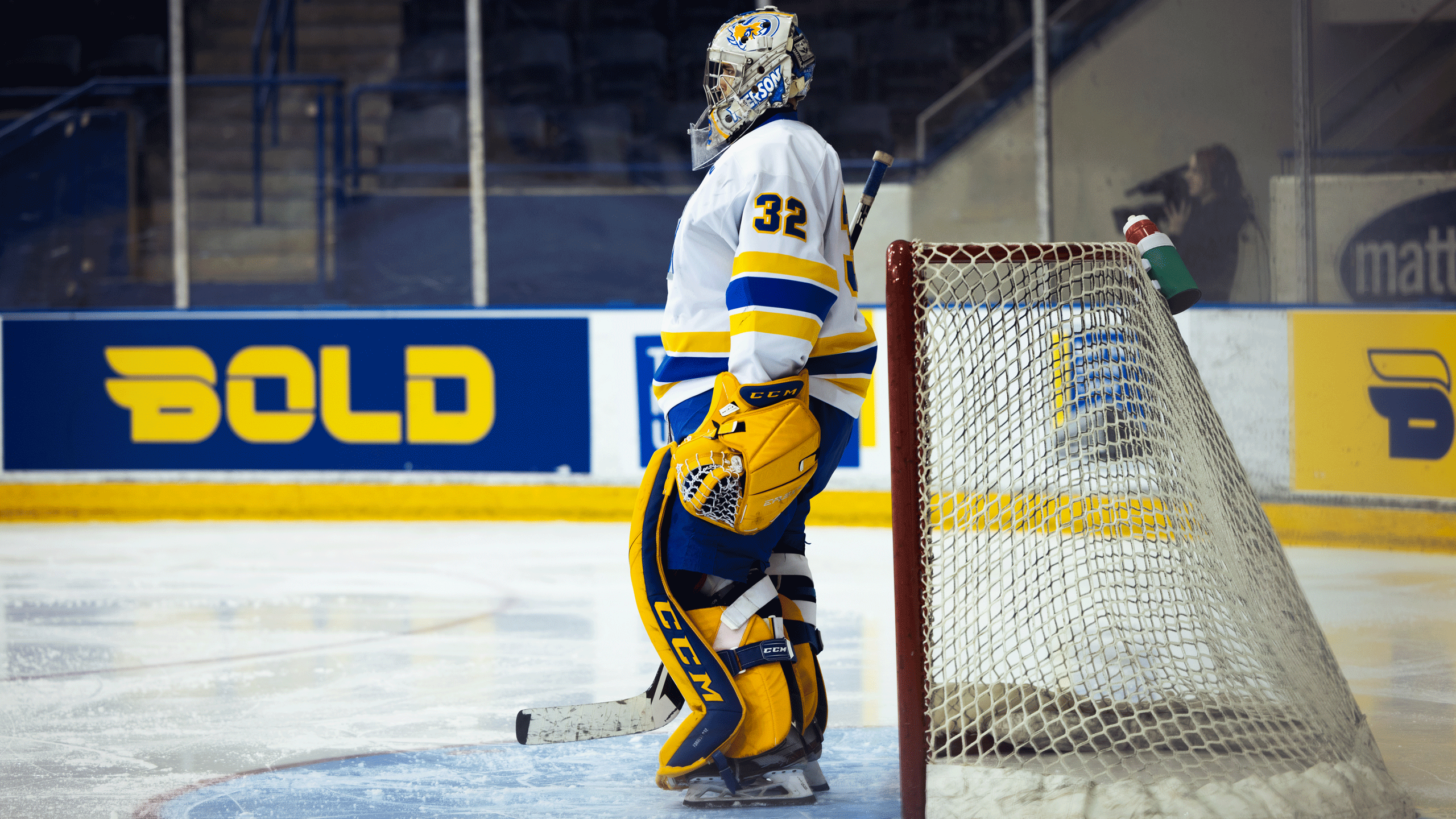 A hockey goalie with yellow pads and a white jersey stands in the crease