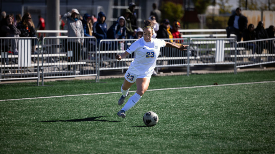A TMU women's soccer player in a white jersey winds up her leg to kick the ball