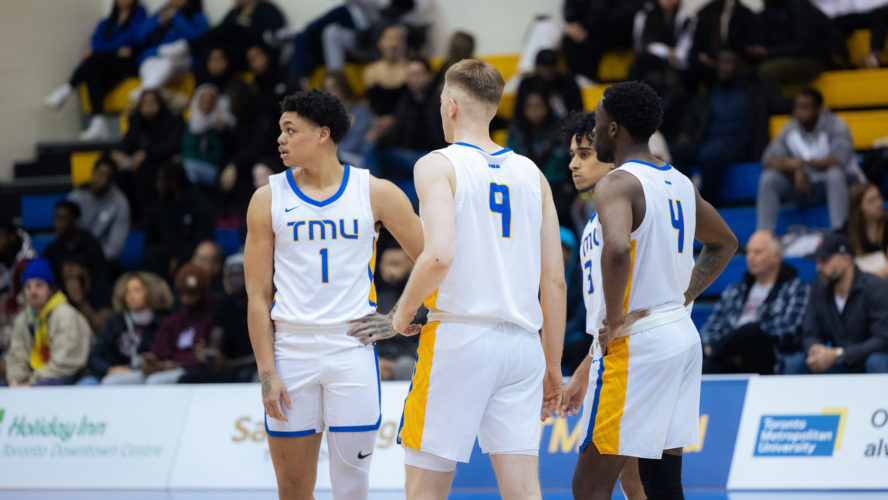 Four TMU men's basketball players in white jerseys deliberate at centre court