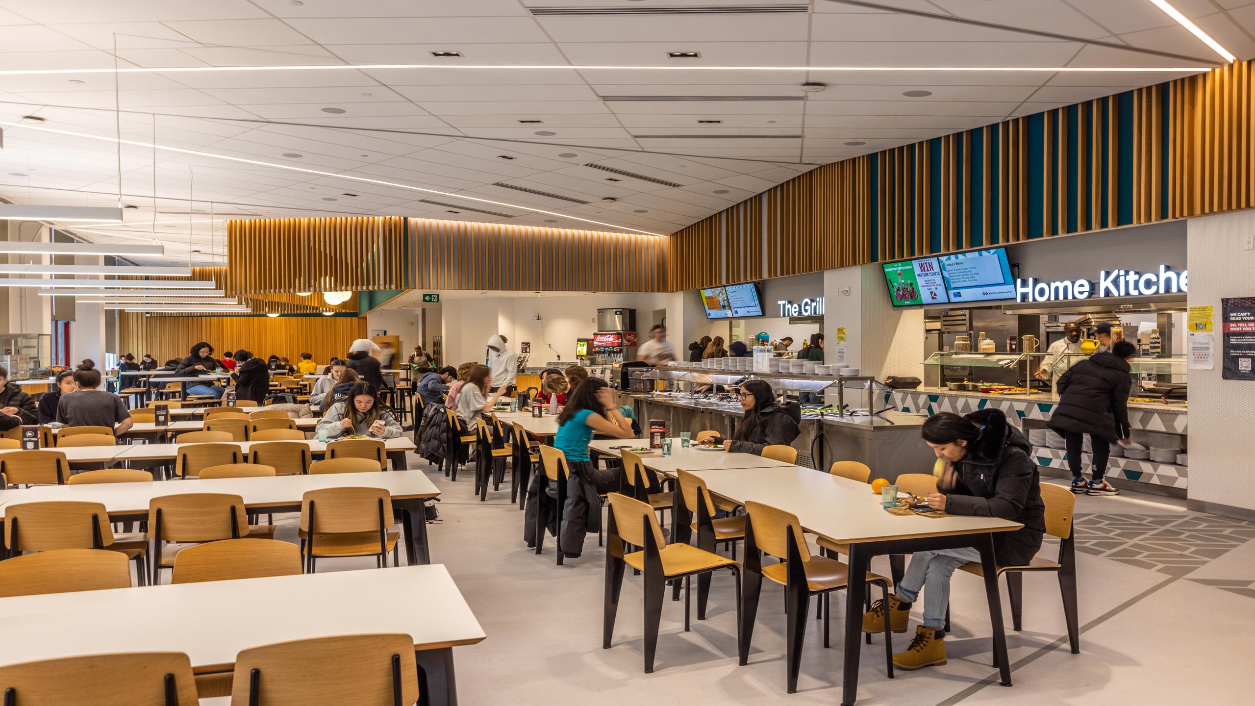 Some students sit at tables in a cafeteria eating while others stand ordering food at a counter