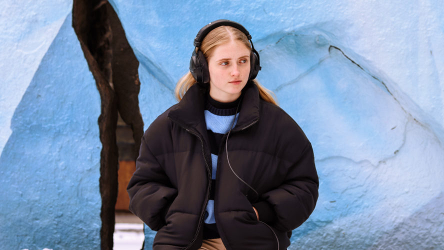 Zoey Ryan pictured wearing a blue sweater and a black puffer jacket with a pair of studio headphones. She sits in front of two rocks painted white and blue to represent glaciers.