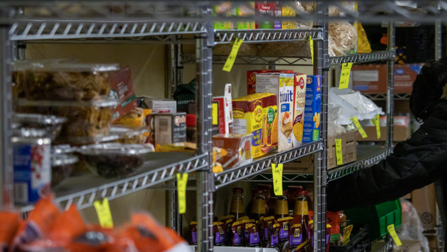 An arm reaches to grab groceries sitting on a metal shelf. There are cereal boxes, trail mix, instant noodles, taco kits and more.