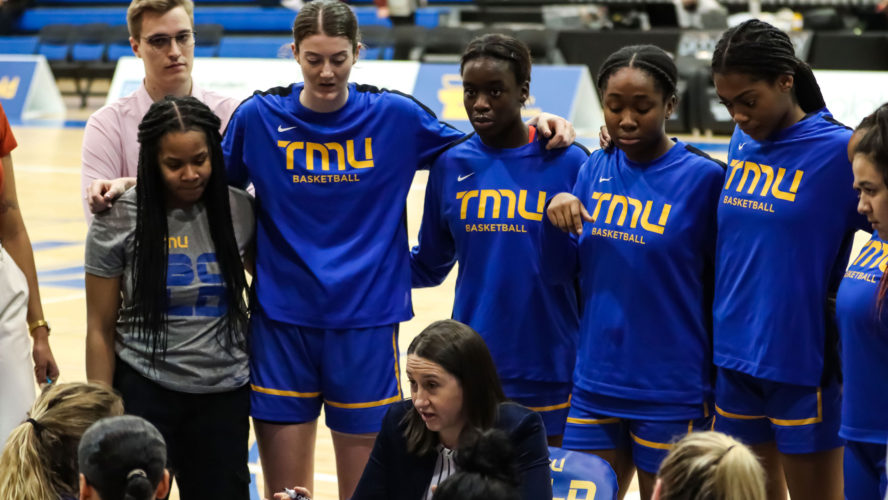 TMU women's basketball players in blue shirts huddle around their coach on the sideline
