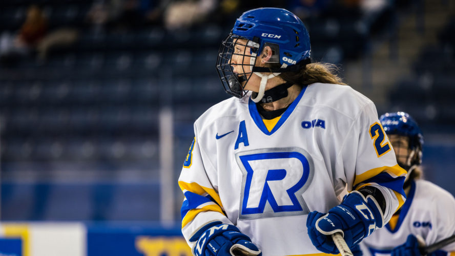 A TMU women's hockey player in a white jersey looks towards the left of the frame towards the puck