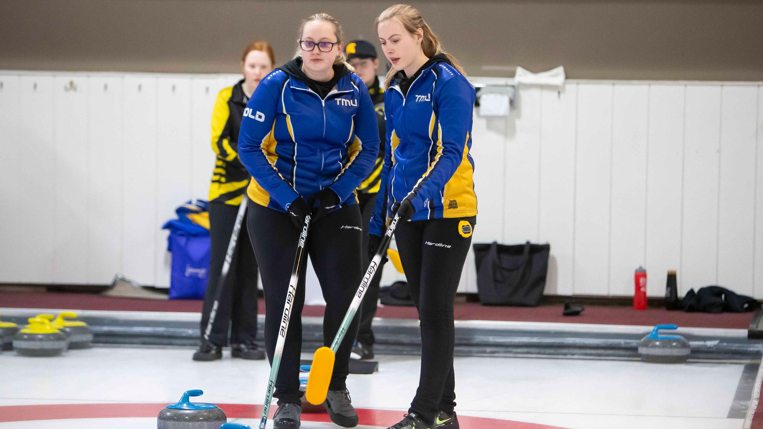 Two TMU women's curlers in blue and gold jackets deliberate in the house about their next shot